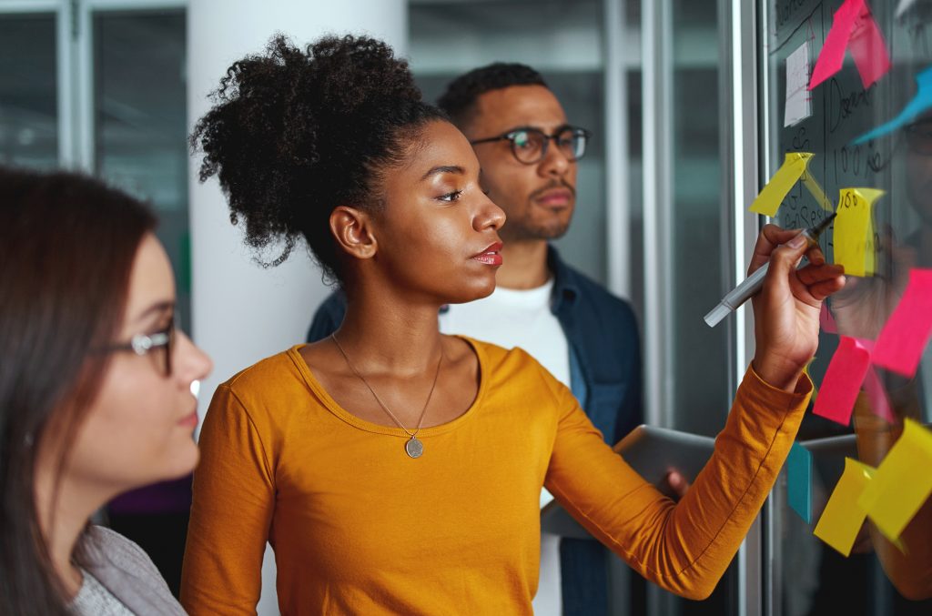 a imagem mostra três pessoas jovens em torno de uma lousa. Uma mulher negra escreve na lousa enquanto outros observam.