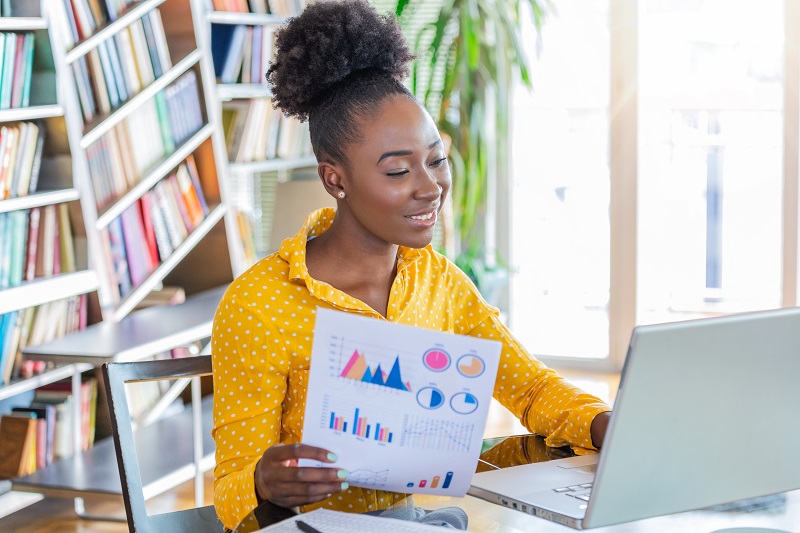 Business professionals. Business woman analyzing data using computer while spending time in the office. Beautiful young grinning professional Black woman in office. Graphs and charts