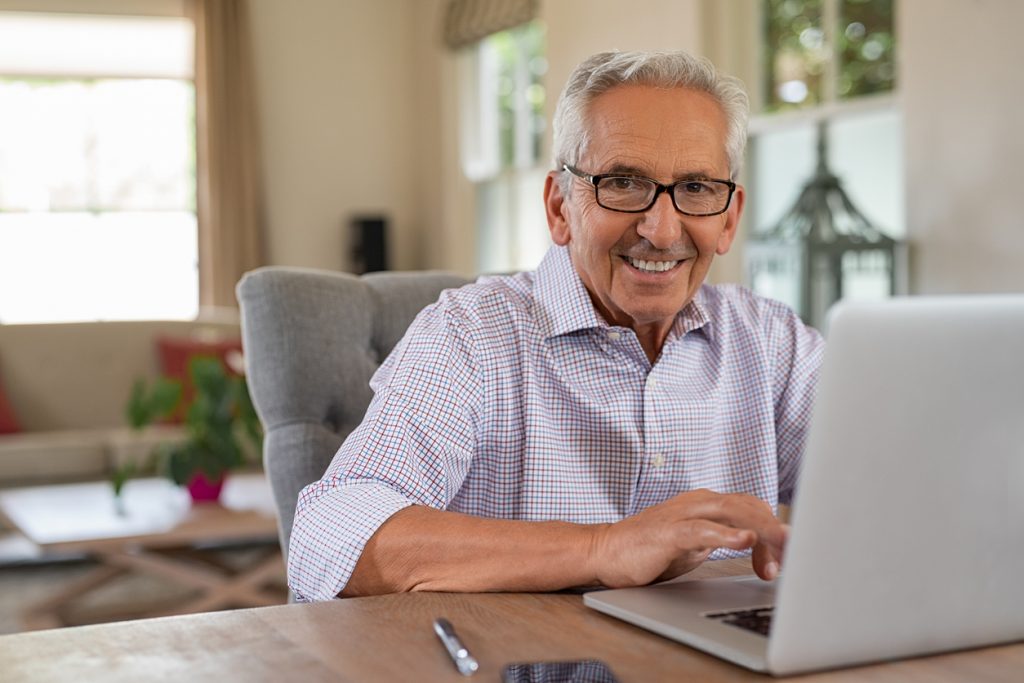 Smiling senior man looking at camera while using laptop at home. Handsome old man wearing eyeglasses working on laptop in living room. Portrait of elderly grandfather using computer.