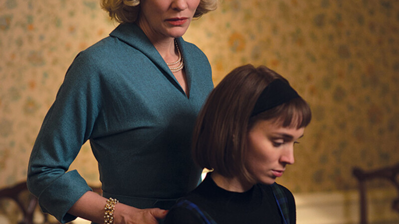 Imagem do filme "Carol". Duas personagens, uma está sentada enquanto a outra encosta por trás com as mãos nos ombros